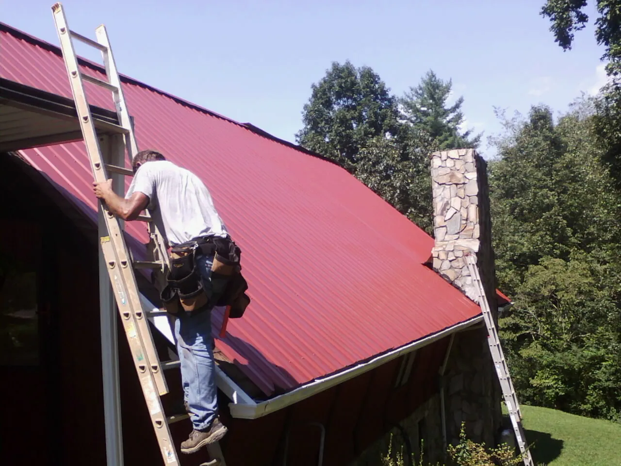 Roofer on a ladder beside a red metal roof and stone chimney — Soffit & Fascia Repair by Roofing Brothers in Racine